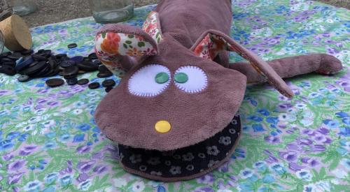 Petite marionnette brune qui bronze sur une nappe à fleurs. Elle a des yeux verts et un petit nez jaune et surtout de grande oreilles colorées. 